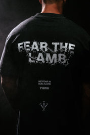 A person shown from the back wearing a black T-shirt with large distressed white text reading 'FEAR THE LAMB' across the upper back and smaller white text below reading 'WE FEAR IN GOD ALONE YHWH'.