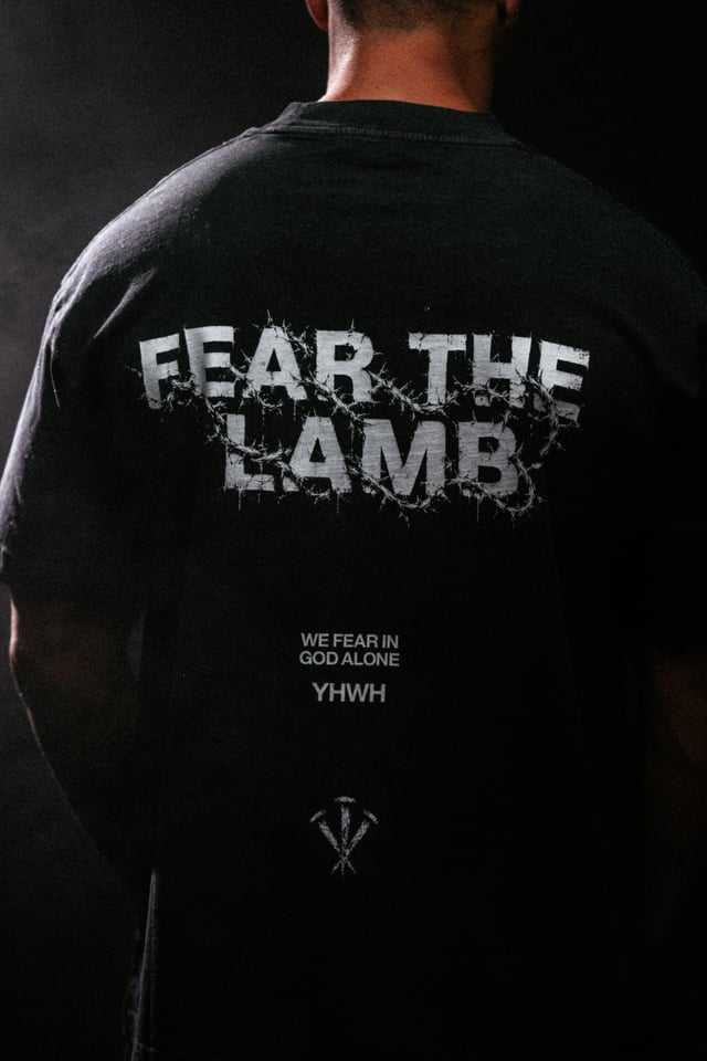 A person shown from the back wearing a black T-shirt with large distressed white text reading 'FEAR THE LAMB' across the upper back and smaller white text below reading 'WE FEAR IN GOD ALONE YHWH'.