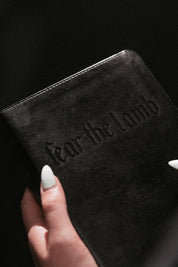Hand holding a book with 'Fear the Lamb' text embossed on the cover against a black background