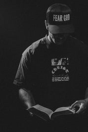 Person wearing a 'Fear God' cap and 'Christ' graphic t-shirt, holding an open book against a dark background