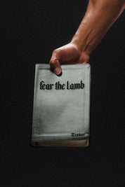 Hand holding a book with 'Fear the Lamb' text embossed on the cover against a black background
