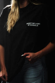 A person wearing a black short-sleeve T-shirt with the white text 'FEAR THE LAMB ETERNALS' printed on the chest, paired with light blue jeans in a dark setting.