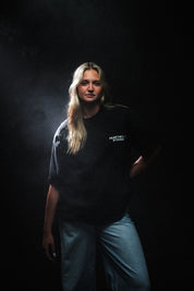 A woman wearing a black short-sleeve T-shirt with the white text 'FEAR THE LAMB ETERNALS' printed on the chest, paired with light blue jeans in a dark setting.