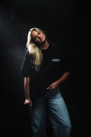A person wearing a black short-sleeve T-shirt with the white text 'FEAR THE LAMB ETERNALS' printed on the chest, paired with light blue jeans in a dark setting.