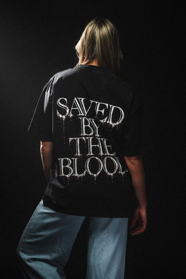 Person wearing a black t-shirt with 'Saved by the Blood' text on the back against a dark background